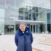 Un prêtre portant manteau, tuque et col romain se tient debout devant la façade de Radio-Canada à Montréal.