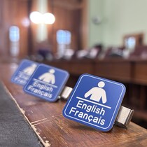 Une photo de la salle des comités parlementaires de l'Assemblée législative. Au premier plan, il y a trois plaques signalant que des services sont disponibles en français et en anglais.