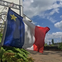 Un drapeau rouge, blanc et bleu avec une étoile jaune dans le coin gauche, planté dans une platebande de verdure devant un église blanche en bois.