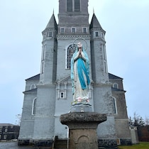 Une église abandonnée et une statue de la Vierge posée sur un socle devant le bâtiment sont photographiées par temps pluvieux, sous un ciel sombre et nuageux.