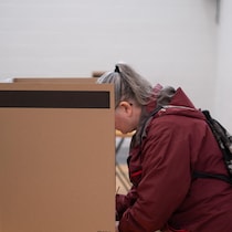 De profil, une femme vêtue d'un manteau bordeaux remplit son bulletin derrière un isoloir en carton.