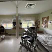 Une petite chambre avec deux lits, deux fenêtres, un rideau au milieu et un fauteuil avec un trou au niveau du siège.