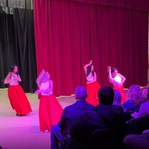 Des danseuses en jupes rouges se produisent sur scène devant un public attentif, sous un éclairage coloré.
