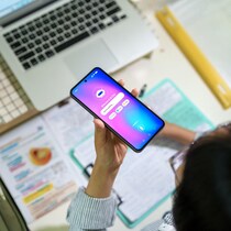 Un adolescent interagit avec une IA sur son smartphone tout en étudiant à son bureau, entouré d'un ordinateur portable, de notes et de fournitures scolaires.