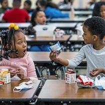 Deux petites filles sont assises dans la cafétéria d'une école pour manger. L'une montre quelque chose qu'elle a dans son lunch et l'autre a la bouche grande ouverte, car elle est émerveillée de ce qu'elle voit.