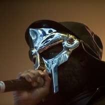 Le chanteur est en prestation sur scène, avec un masque argent et une cape. Il tient un micro.