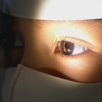 Gros plan sur le visage d'un enfant qui passe un examen de la vue. Une lumière éclaire l'un de ses yeux et il porte un masque.