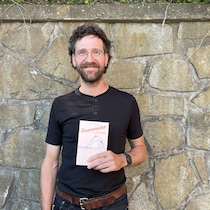 Un homme tenant un livre dans sa main. Il est à l'extérieur devant un mur de pierres.
