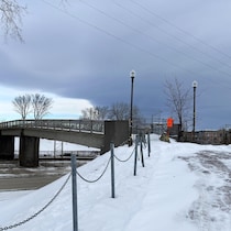 Vue en contre-plongée d'une structure de pont en béton gris traversant une route dégagée. Le sol est recouvert d'une épaisse couche de neige blanche et de glace. Au premier plan, une clôture faite de poteaux métalliques reliés par des chaînes noires borde un sentier glacé. Le ciel est couvert de nuages sombres et menaçants.