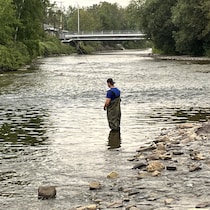 Un homme pêche debout dans une rivière, tandis qu'on voit un petit pont au loin.