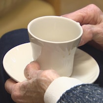 Une personne âgée, dont on ne voit que les mains, boit un café d'une tasse en procelaine.