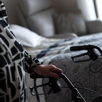 Une personne âgée, dont on voit la main posée sur un déambulateur, à côté d'un lit.