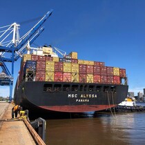Un navire sur lequel sont empilés de deux à trois étages de conteneurs de marchandise de différentes couleurs est amarré à un quai, sous un ciel bleu.