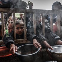 Des enfants tendent des bols entre les barreaux d'une clôture.