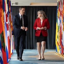 S'avançant vers la caméra, Rob Lantz et Susan Holt marchent entre deux rangées de grands drapeaux des provinces. Il parle et elle tourne la tête vers lui.
