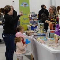 Une femme et deux jeunes enfants devant une table où s'empilent des dizaines de livres. La femme est en conversation avec un des deux personnes derrière la table. 