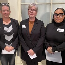 Trois femmes d'âges divers se tiennent droites, les mains jointes en tenant des documents.