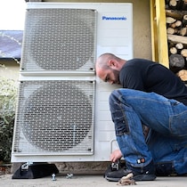 Un travailleur photographié de profil, un genou au sol, en train d'installer une thermopompe sur le côté d'une maison, à côté d'une pile de bûches.