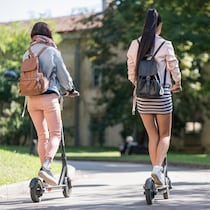 Deux femmes vues de dos, avec de petits sacs à dos, circulent sur des trottinettes électriques et s'apprêtent à prendre une courbe légère dans une allée bordée d'arbres feuillus. 