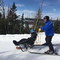 Jean-François Rioux, chaussé de skis, pousse sur une piste une personne dans une chaise adaptée.