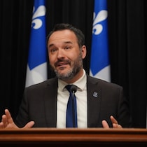 Le ministre s'exprime devant des drapeaux du Québec.