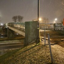 Photographie de nuit sous une légère neige, montrant une passerelle cyclo-piétonne en béton et acier enjambant une autoroute. L'accès à la passerelle au premier plan est barré par des clôtures temporaires et un panneau de signalisation orange triangulaire indiquant "Trottoir Barré". Du ruban jaune et noir, similaire à celui de la police (crime scene tape), est enroulé autour d'un poteau et s'étend sur le sol. La scène est éclairée par des lampadaires.