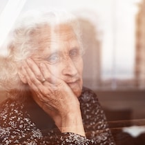 Une dame âgée, la tête dans sa main, regarde par la fenêtre.