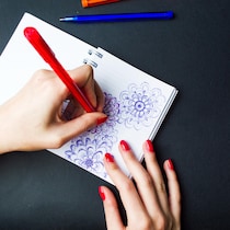 Gros plan sur des mains de femme dont celle de gauche dessine des fleurs dans un cahier.