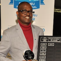 Jean Claude Ngendahayo, président de l'ANFSO-ESPOIR est debout devant une affiche du Club Richelieu et tient dans ses mains une plaque et un médaillon qui font partie du prix de l'Organisme novateur de l'année 2023 - 2024 du club Richelieu de Windsor.