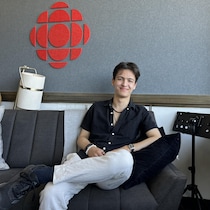Un homme souriant est assis sur un sofa avec derrière lui, le logo de Radio-Canada.