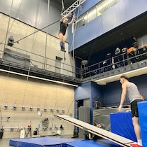 Une jeune femme athlétique est prise en photo en plein vol dans les airs, dans une grande salle d'entraînement, après avoir été propulsée par une planche coréenne qui fonctionne comme une balançoire à bascule.