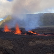 Image d'un sol volcanique plutôt plat en éruption incandescente. Au loin on distingue des montagnes sous un ciel ensoleillé.