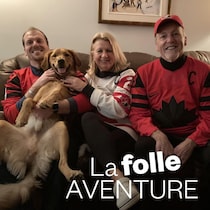 Trois personnes portant des chandails de hockey de l'équipe canadienne sont assises dans un divan avec un chien.