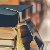 Un mortier, un ordinateur portable, un diplôme et des livres posés sur une table dans une bibliothèque.
