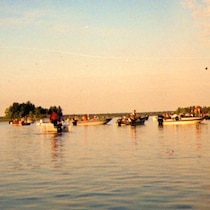 Plusieurs bateaux et leurs passagers, rassemblés sur un lac, à l’aube.