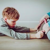 Un parent assis par terre essaie d'enlever des mains de son enfant allongé sur le sol sa tablette qu'il retient avec ses deux mains. 
