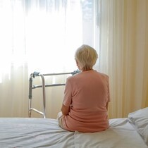 Vue de dos, une femme âgée est assise sur un lit dans une chambre.