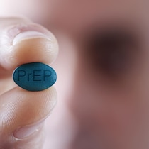 Photo rapprochée d'une personne qui tient une gélule du médicament PrEP qui est inscrit dessus. On voit une partie du visage flou de la personne au second plan