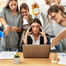 Un groupe d'employés crient vers une personne stressée qui a les yeux fermés et essaie de respirer en se tenant la tête dans les mains devant son ordinateur portable.
