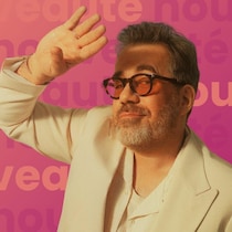 Un homme avec une barbe grise et des lunettes de soleil teintées orange pose avec une main levée pour se protéger les yeux du soleil. Il porte une veste de tailleur crème décontractée et une chemise en maille. L'arrière-plan montre une route en montée bordée d'arbres à Los Angeles, en Californie.