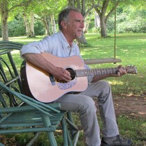 Zachary Richard est assis sur un banc dehors et il joue de la guitare.