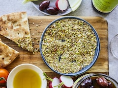 Un bol de dukka aux pistaches et aux amandes sur une planche en bois avec des légumes et du pain autour. 