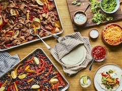 Une plaque de porc et de légumes ainsi qu’une plaque de haricots noirs et de légumes sont accompagnées d’une assiette de petites tortillas de blé, d’un bol de salsa, d’un bol de fromage, d’un bol de coriandre et d’assiettes avec des fajitas.