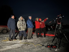 5 hommes regardent le ciel en soirée. L'un d'eux pointe le ciel avec son doigt. En avant-plan se trouve un télescope.