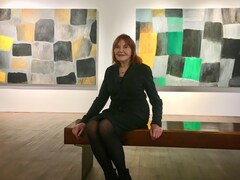 La femme est assise sur un blanc, placé devant ses toiles, et elle sourit. 