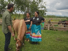 Un cheval broute du gazon pendant que deux femmes autochtones discutent avec un homme.