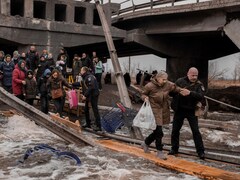 Des gens traversent une rivière sur une planche de bois sous un pont détruit.