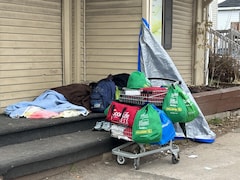 Une personne itinérante allongé sur les marches d'un logement. Un chariot est rempli de sacs qui contiennent ses affaires. 