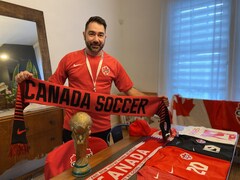 Un homme portant un gilet de l'équipe canadienne de soccer, tenant un foulard rouge où est inscrit «Canada soccer». Divers articles représentant l'équipe se trouvent devant lui sur une table.