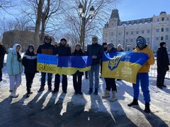 Un groupe de personne devant l'Assemblée nationale. Certains tiennent devant eux un drapeu ukrainien déplié. Le ciel est bleu et il y a de la neige au sol.